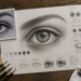 Estudo artístico em grafite de um olho realista, com fotografia de referência, estudos da íris, pálpebras, luz e sombra e materiais de desenho sobre a mesa